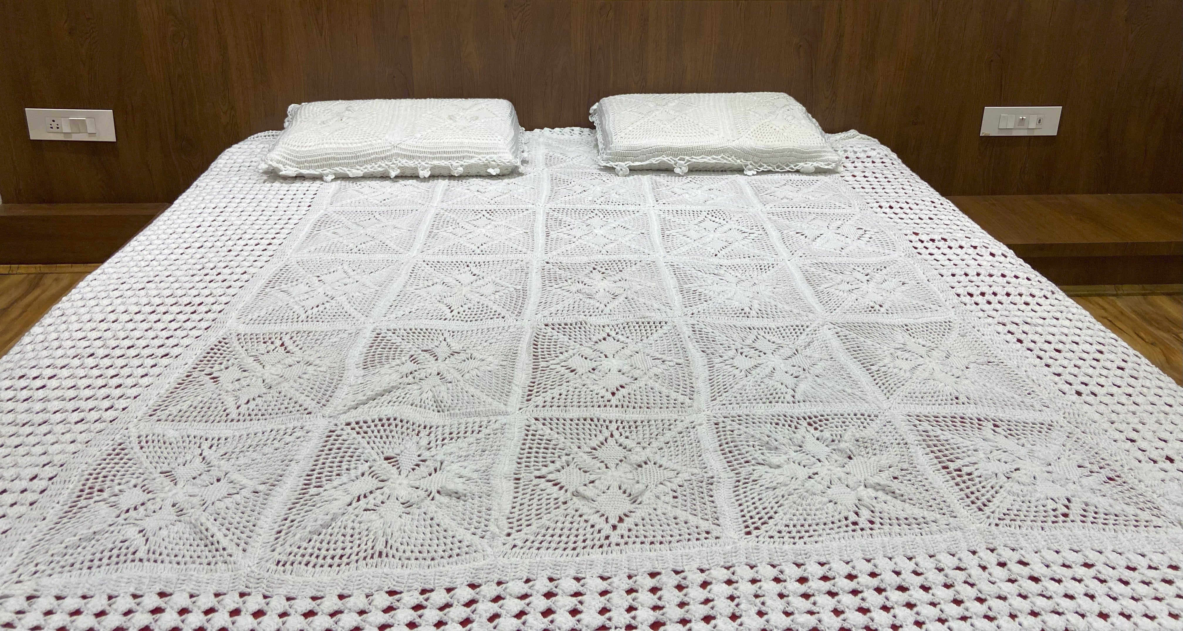 Bed with a white crocheted blanket and two pillows against a wooden wall.